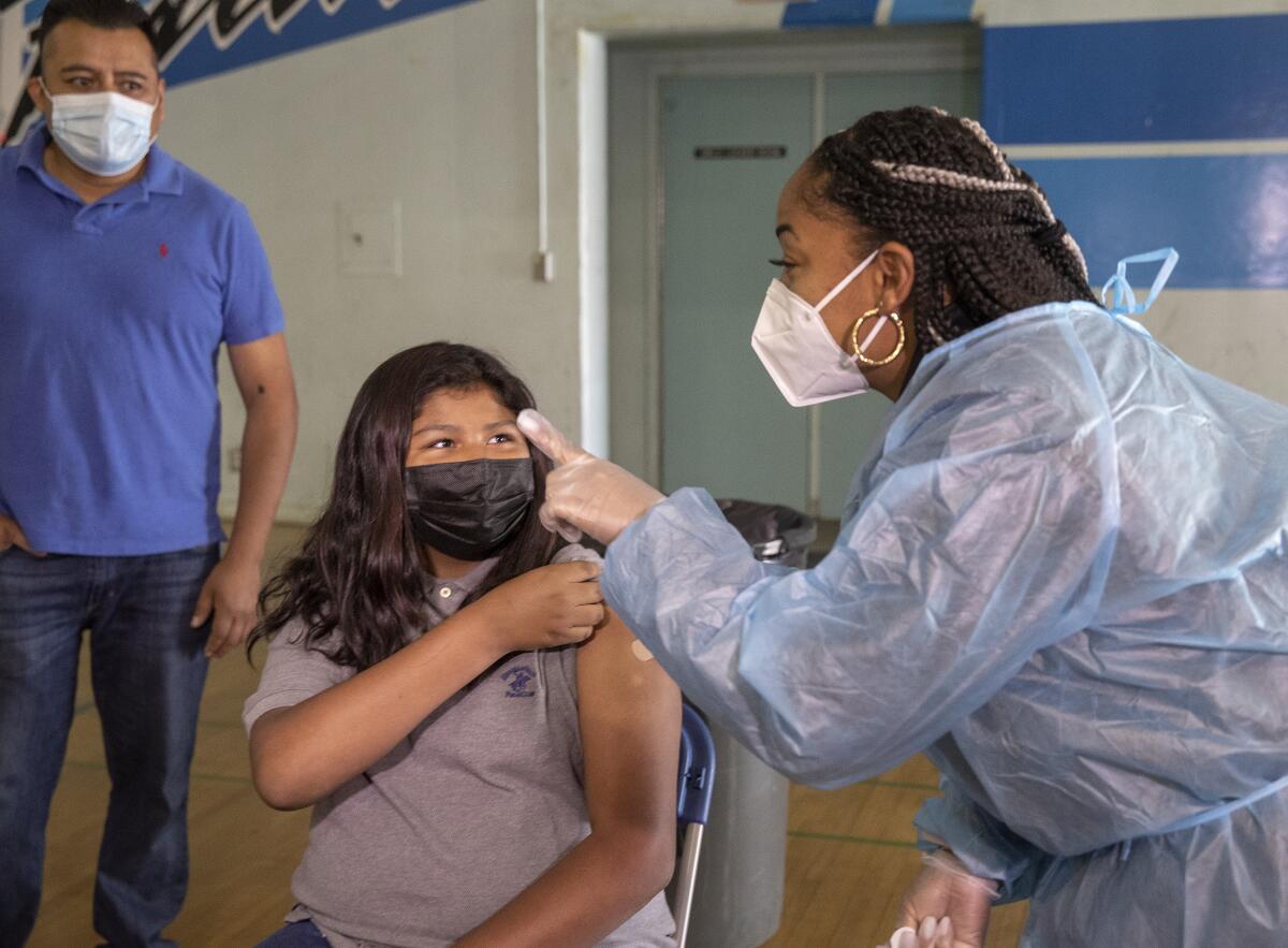 A girl rolls up a sleeve as a person in a face mask and body covering points a finger. 