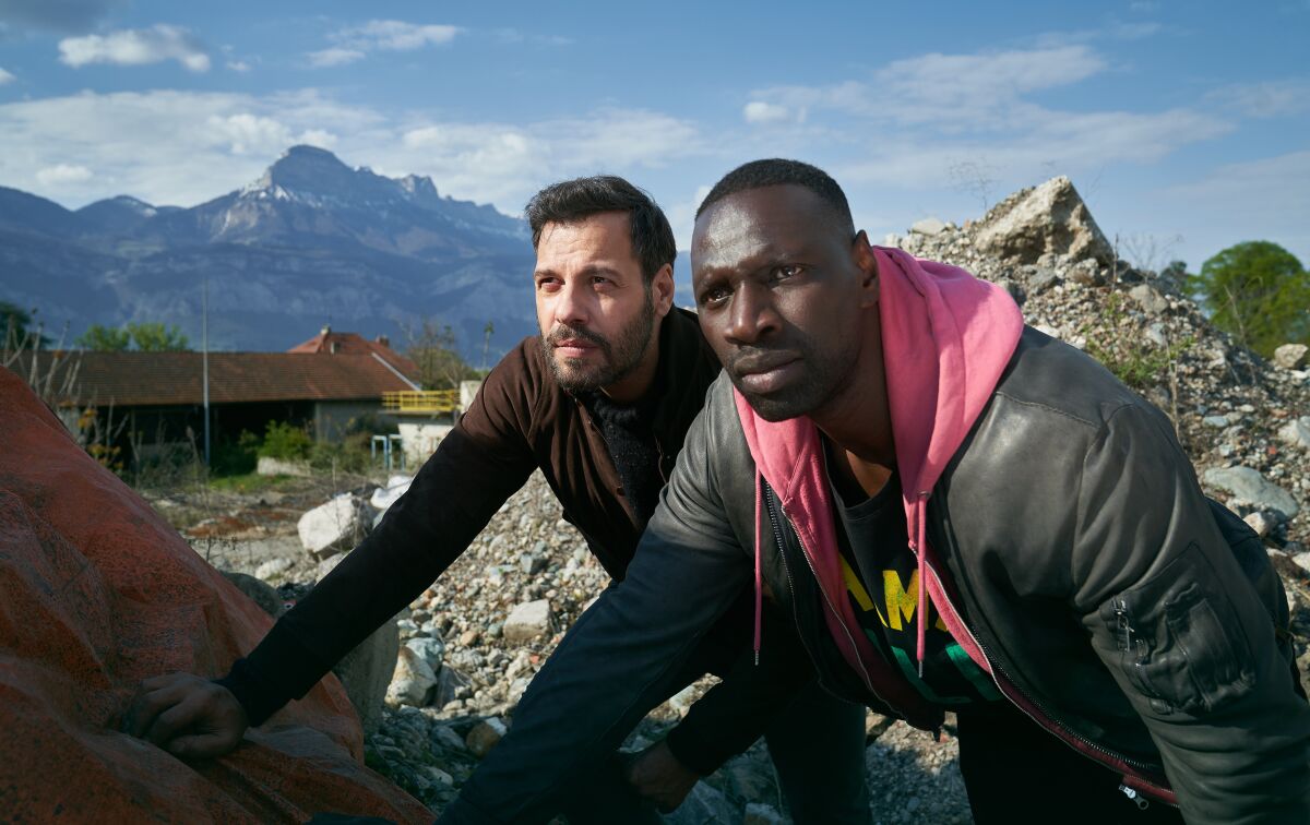 Two men lean on a rock against a mountainous backdrop in the movie "The Takedown."