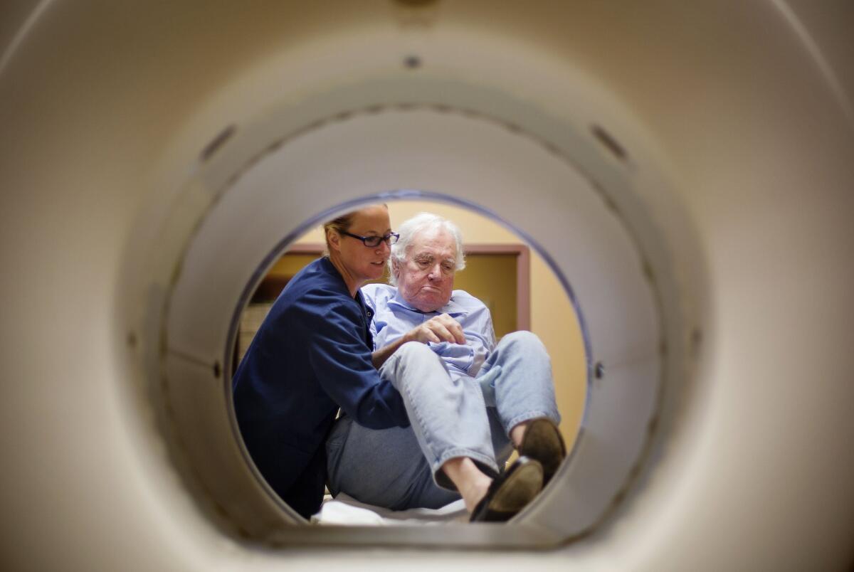 A man in is helped off a table by a technician after undergoing a CT scan.