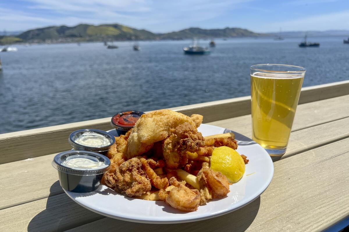A plate of food and a glass of beer on a wooden counter overlooking the ocean, with hills in the distance