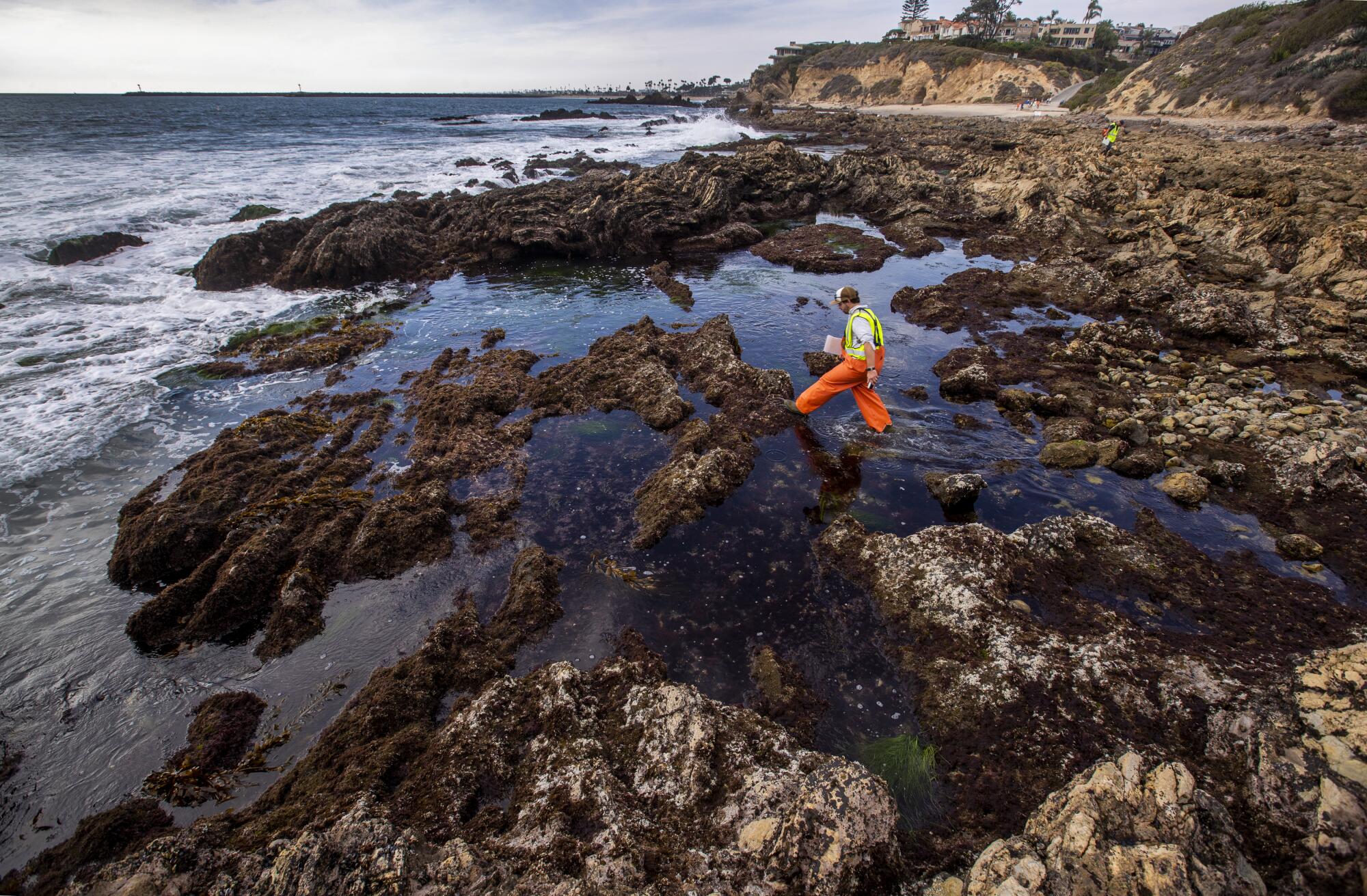 A man in red coveralls walks among rocks on the shore 