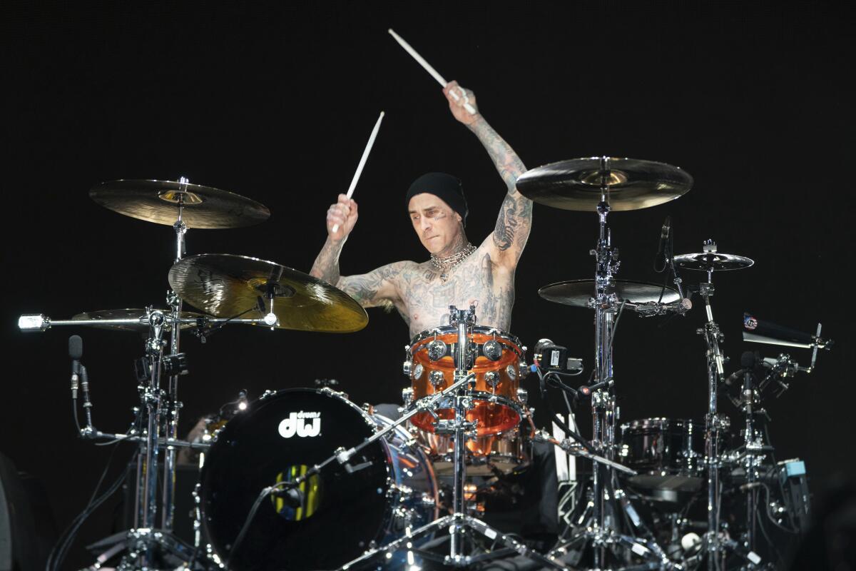 Travis Barker wears a black beanie and no shirt as he holds drum sticks above his head and an orange drum set onstage