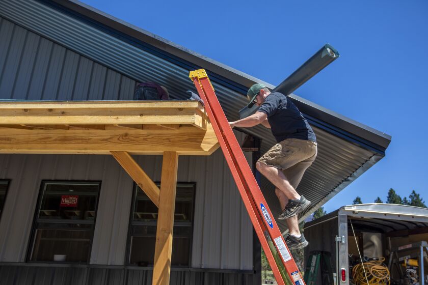 Greenville, CA - July 15: Kaley Bentz, owner of Riley's Jerky carries roofing tile up a ladder on Friday, July 15, 2022, in Greenville, CA. His family's beef jerky business burned down in the Dixie Fire one year ago. He is in the process of rebuilding. (Francine Orr / Los Angeles Times)