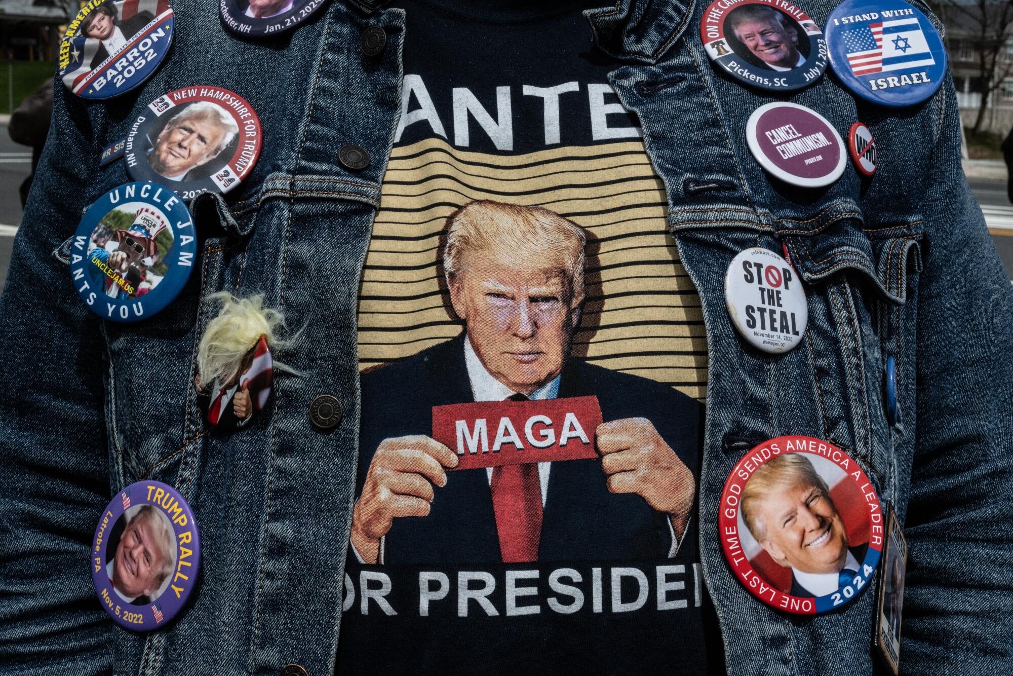 O peito de uma pessoa vestindo uma camisa Trump e uma jaqueta jeans decorada com 12 botões políticos, a maioria deles para Trump.