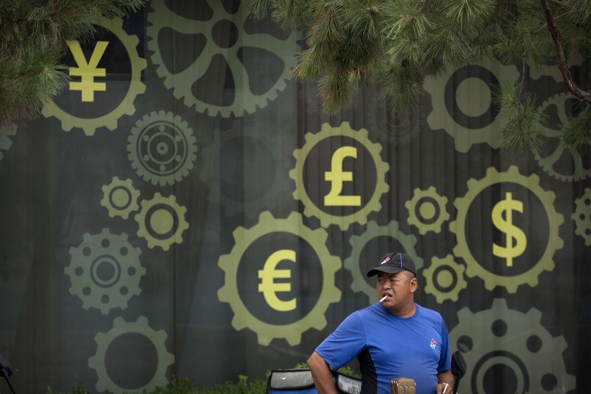 FILE - In this July 20, 2018, file photo, a deliveryman stands near a mural displaying Chinese yuan and other world currency symbols on the outside of a bank in Beijing. The Tokyo Olympics are already the most expensive Summer Games on record with costs set to go higher, a wide-ranging study from Britain's University of Oxford indicates. This is even before the costs of the one-year delay from the COVID-19 pandemic are known with the cost overrun already at over 200%, lead author Bent Flyvbjerg explained in an interview with the Associated Press. (AP Photo/Mark Schiefelbein, File)