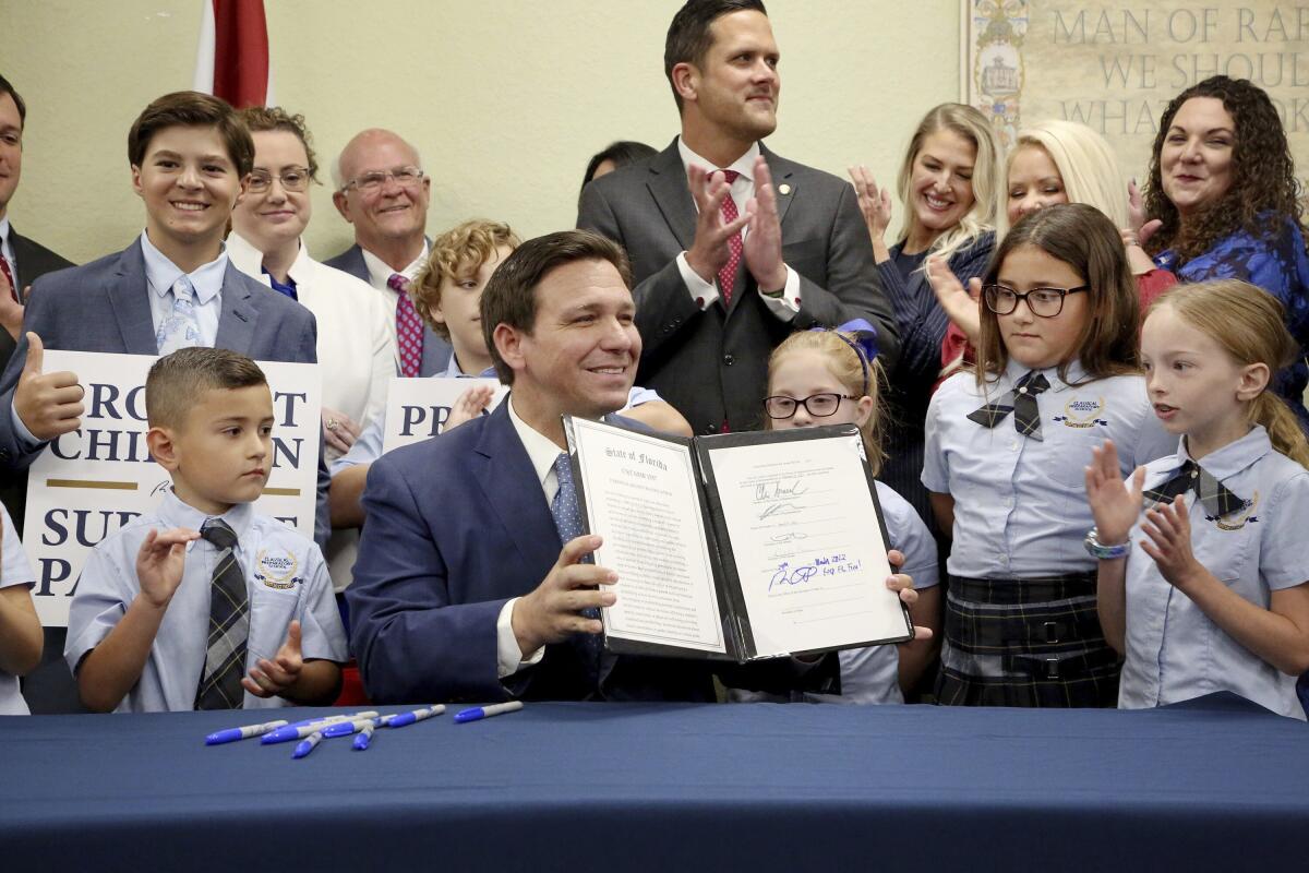 Flanked by children in uniforms, Florida Gov. Ron DeSantis holds up a signed document as adults smile and applaud behind him.