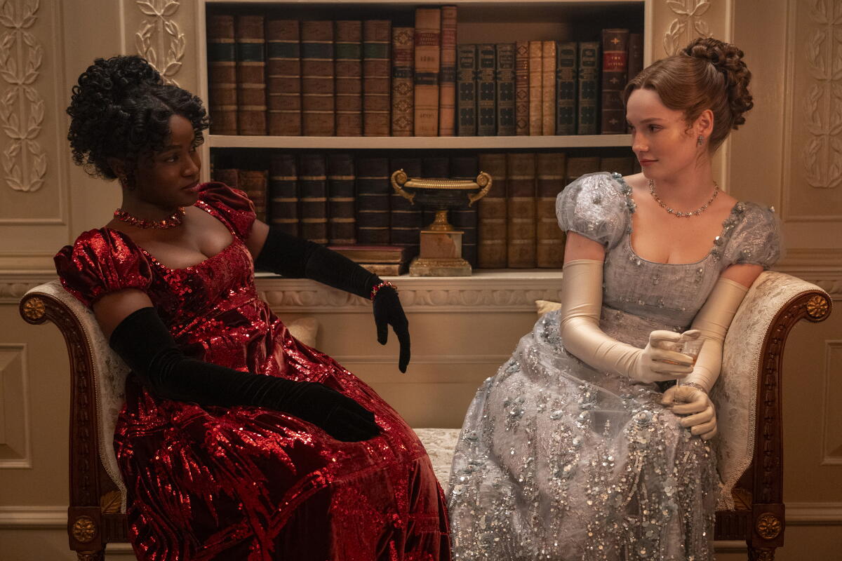 Two women sit on a settee in period gowns and gaze at each other