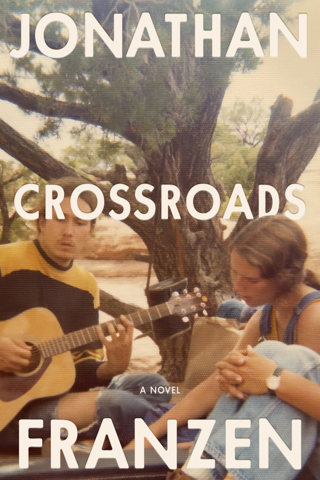 A man playing the guitar and a woman are sitting outside on the blanket of "Crossroads," by Jonathan Franzen.
