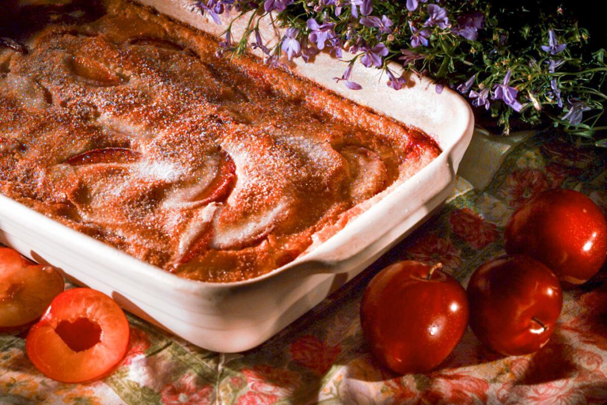 A plum clafoutis in a white dish is surrounded by plums and purple flowers.