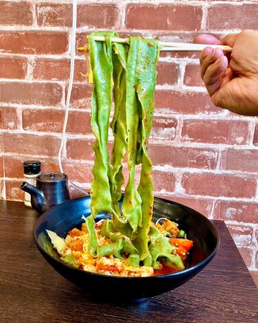 Chopsticks lift wide green noodles from a black bowl filled with vegetables and more noodles