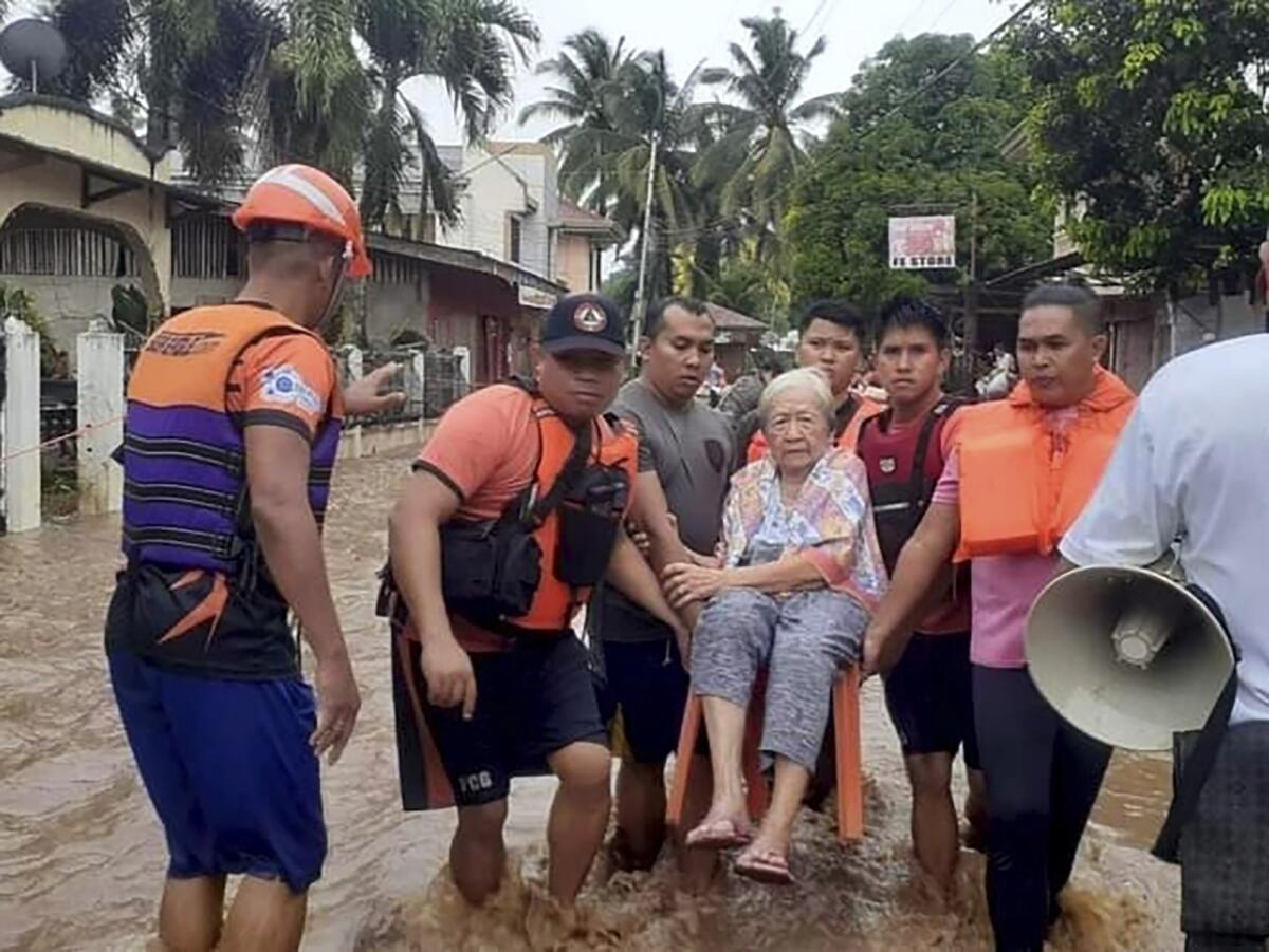 An elderly woman sits on a chair while being carried by coast guard personnel through floodwaters.