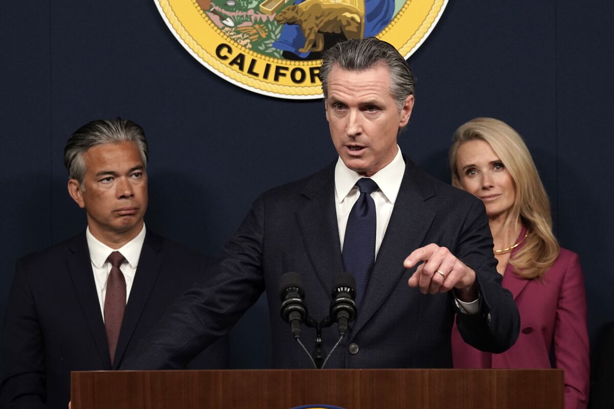 California Gov. Gavin Newsom points while talking at a lectern and a man and woman stand behind him on either side.