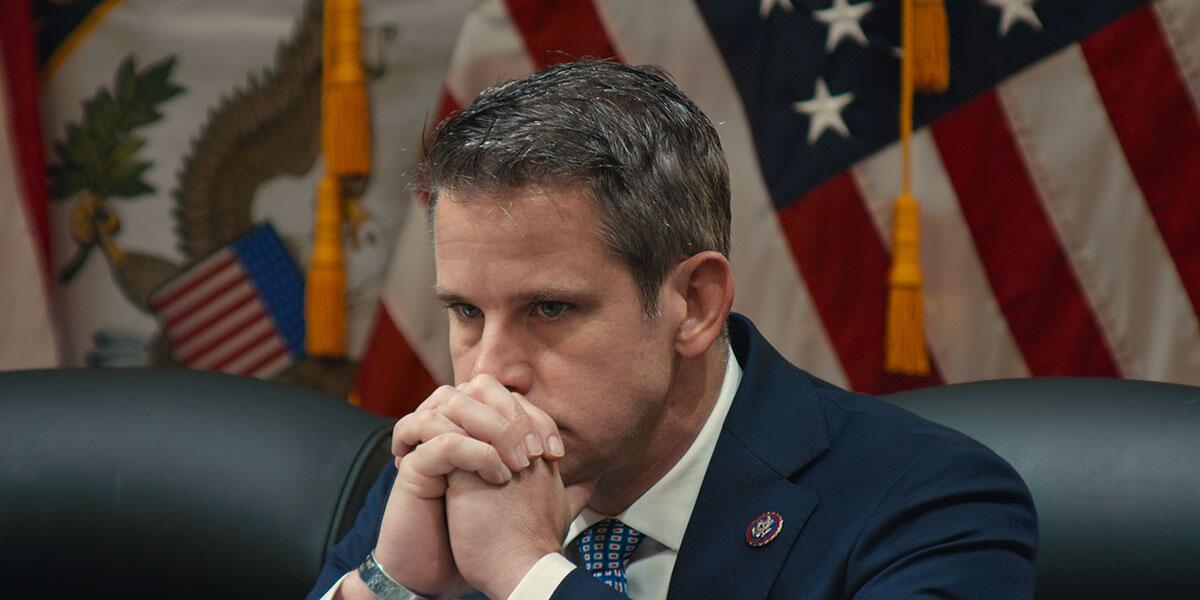 A congressman with flags behind him holds his hands to his face.