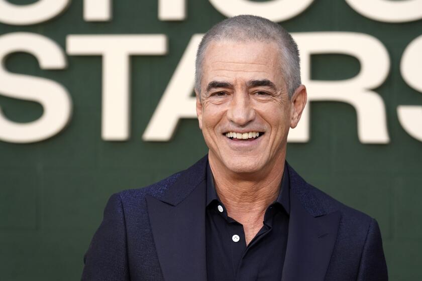 Dermot Mulroney with buzzed hair in a blue blazer and dress shirt smiling against a green backdrop