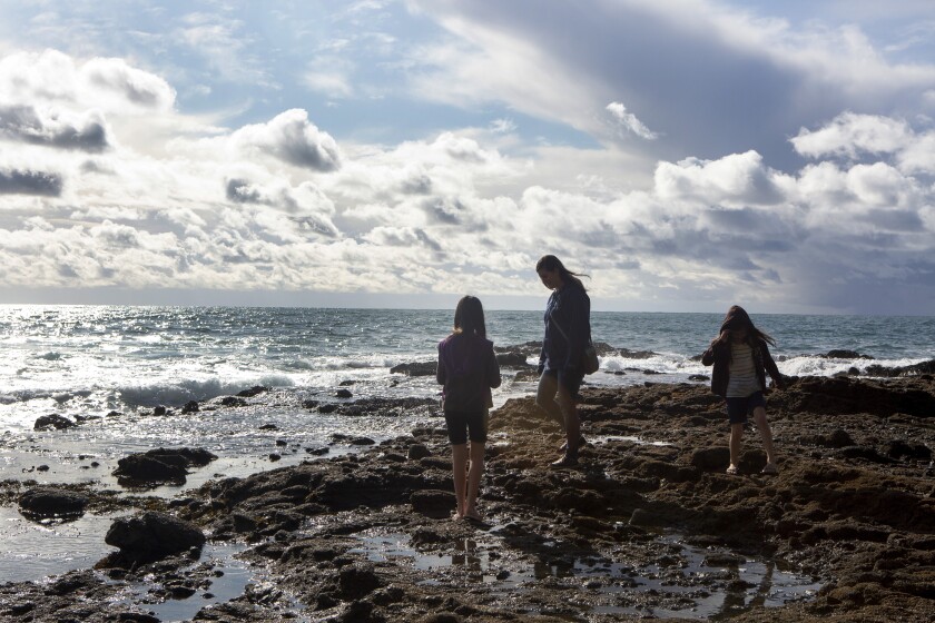 Three young people stand on a rocky shoreline as the sun glints off rolling ocean waves.