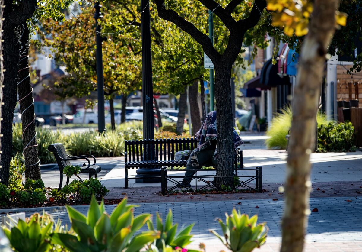 A homeless man sits under a blanket on a bench