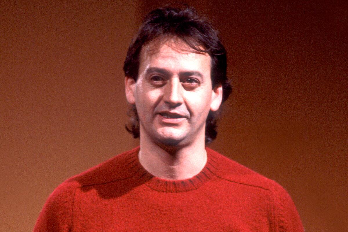 Comedian Joe Flaherty, sporting a red sweater and mullet, performs on stage.