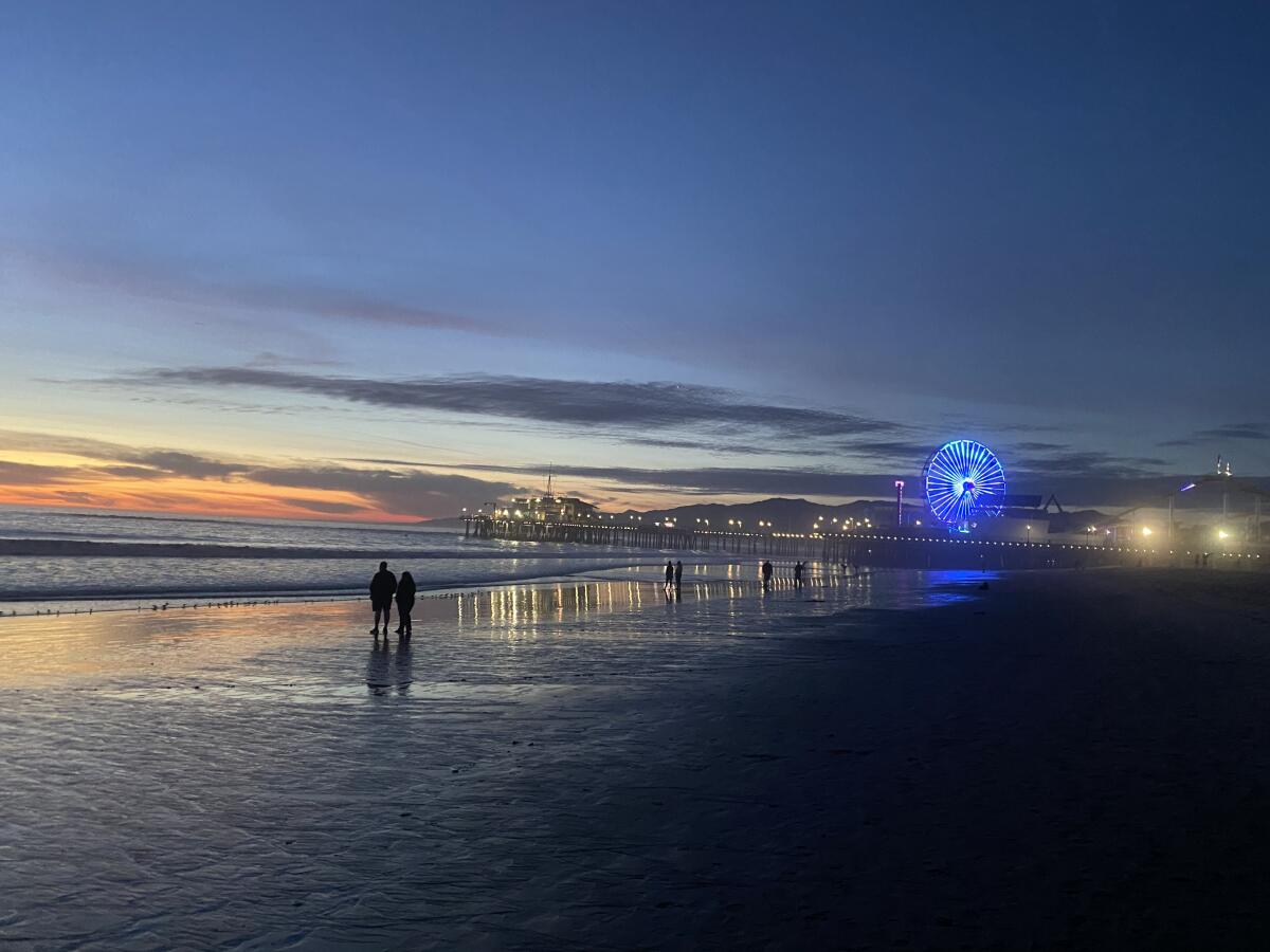 Two people walk along the beach as a ferris wheel lights up in the distance