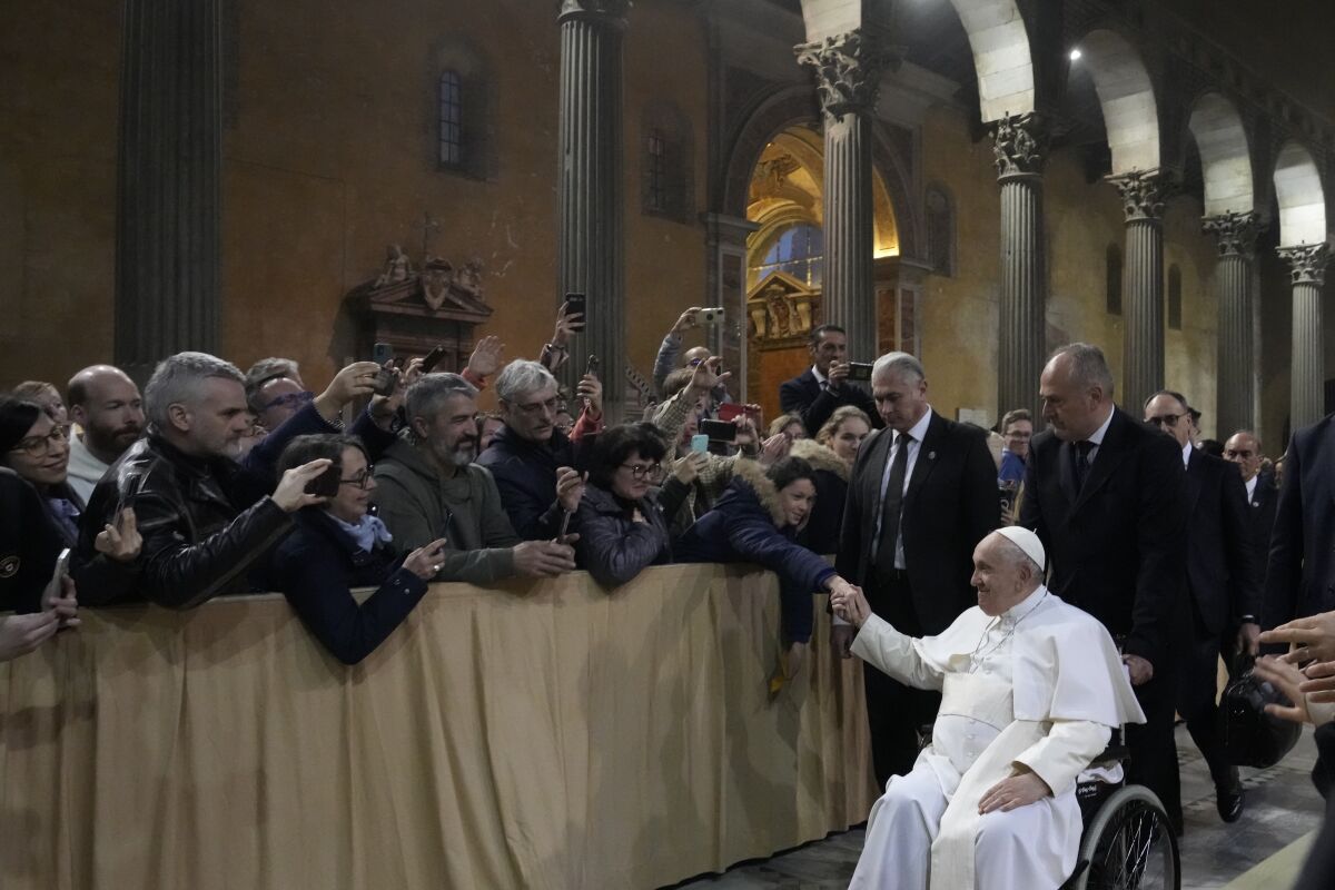 Pope Francis, using a wheelchair, reaching up to shake hands with people behind a barricade
