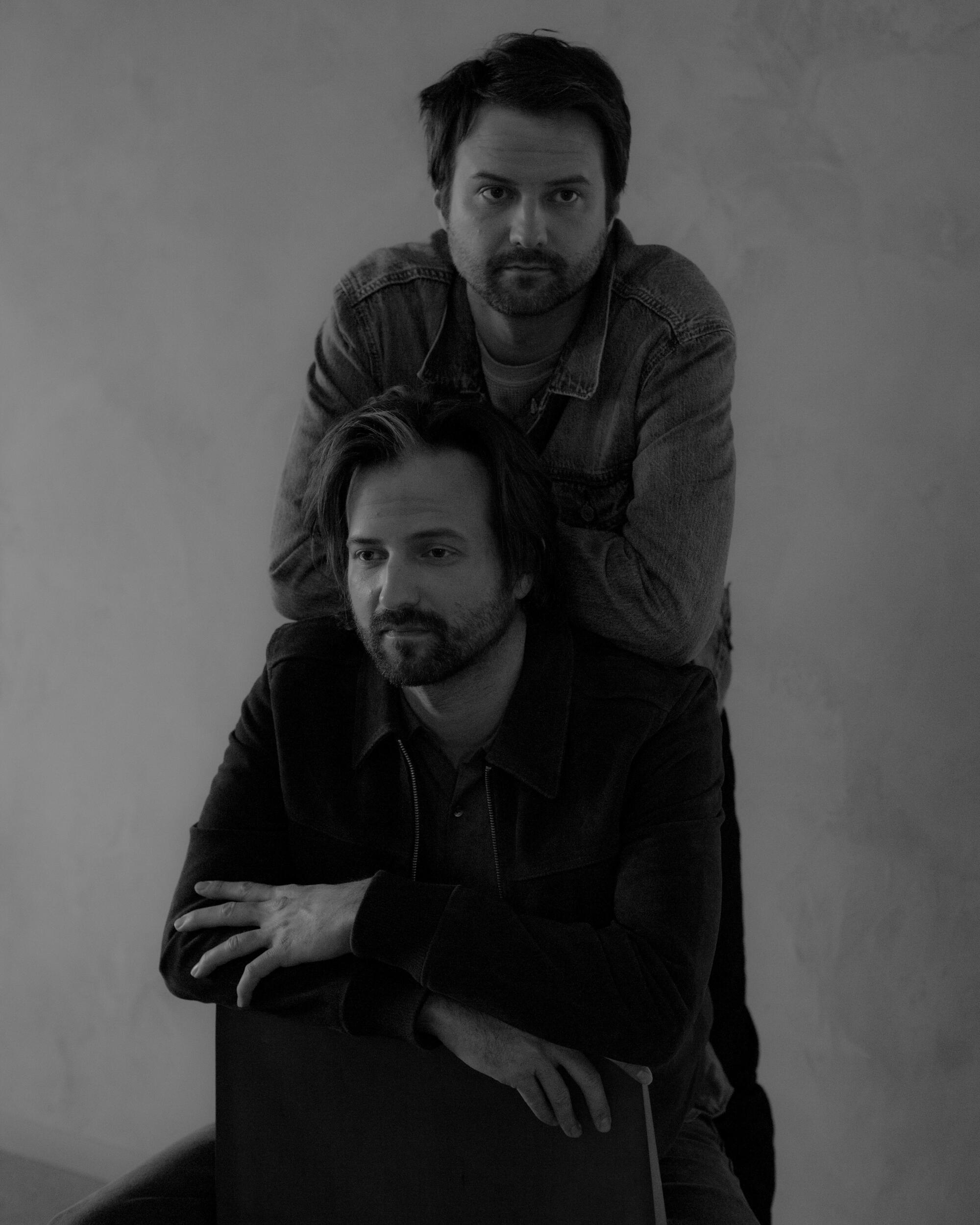 Two men pose for a portrait, one seated and the other leaning on his back