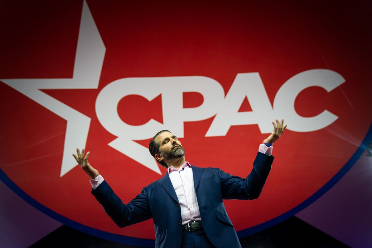 Donald Trump Jr. looks to the sky with hands spread in front of a large CPAC logo