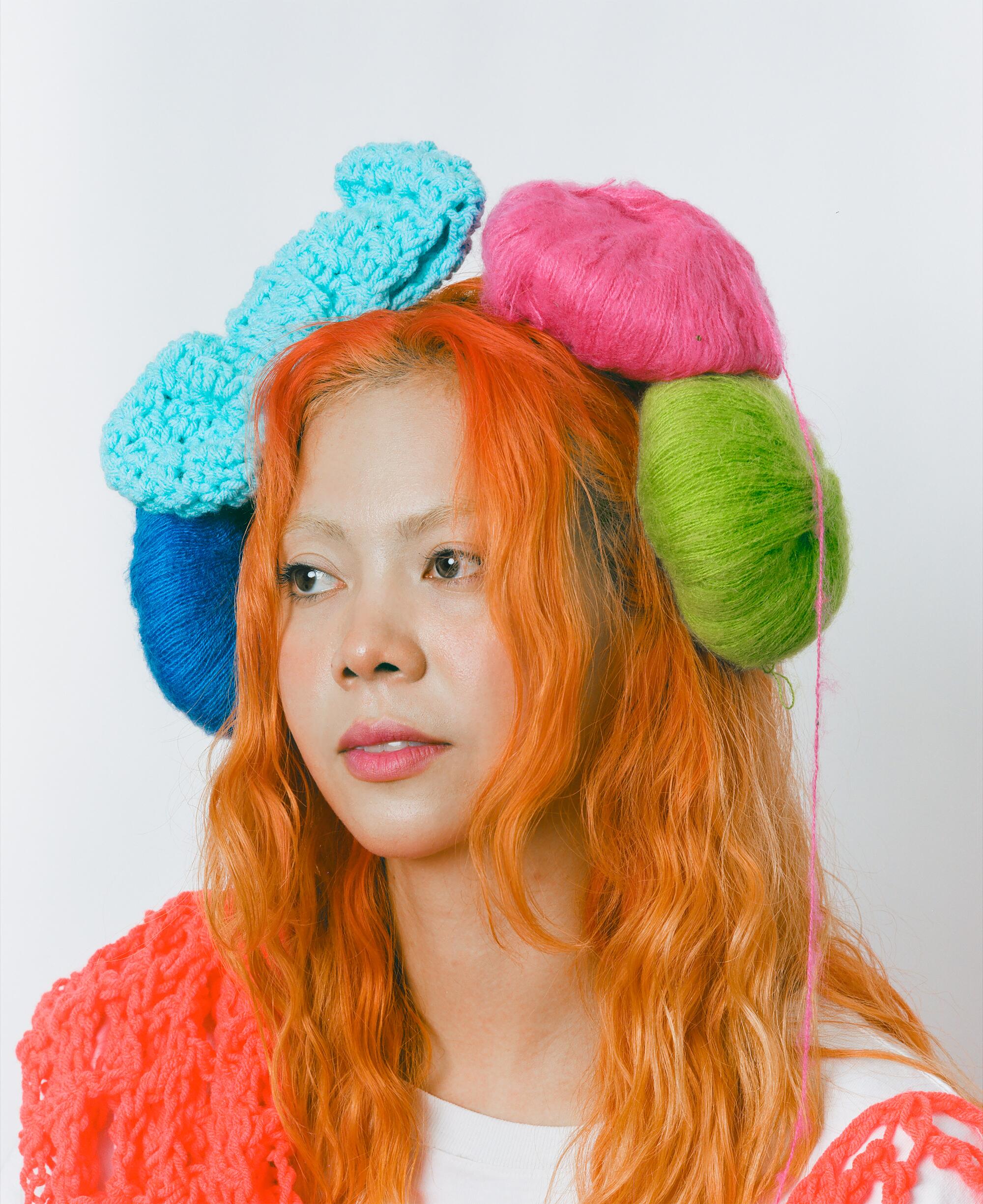 A woman with orange hair wears colorful balls of yarn as a headpiece.