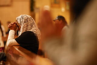A woman wearing a white lace veil kneels in a church pew.