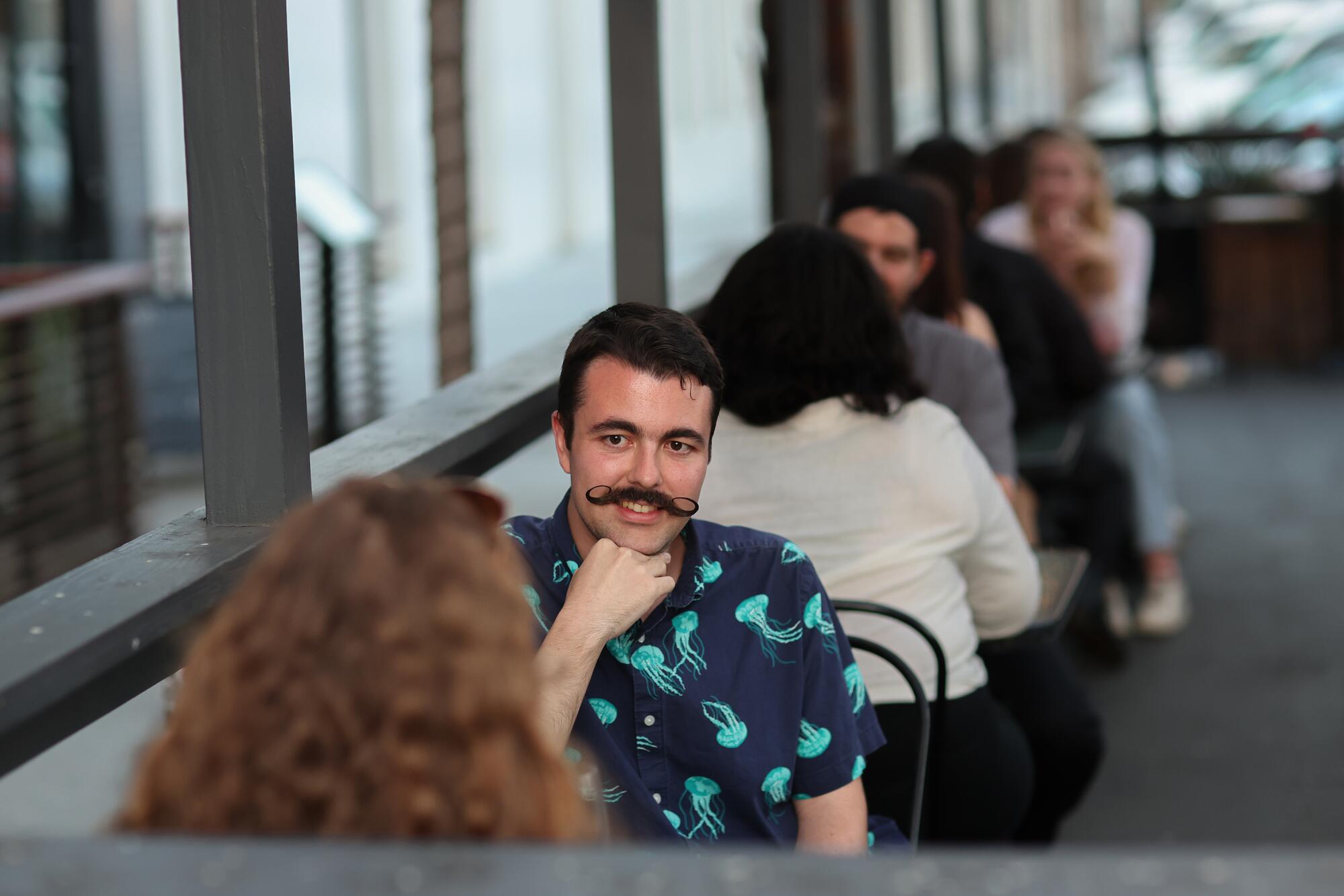 A mustached man facing the camera talks to a woman at a speed dating event.