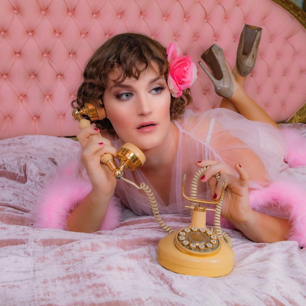 A woman in a pink nightdress with a flower in her hair lines on her stomach in a bed with a rotary phone to her ear 