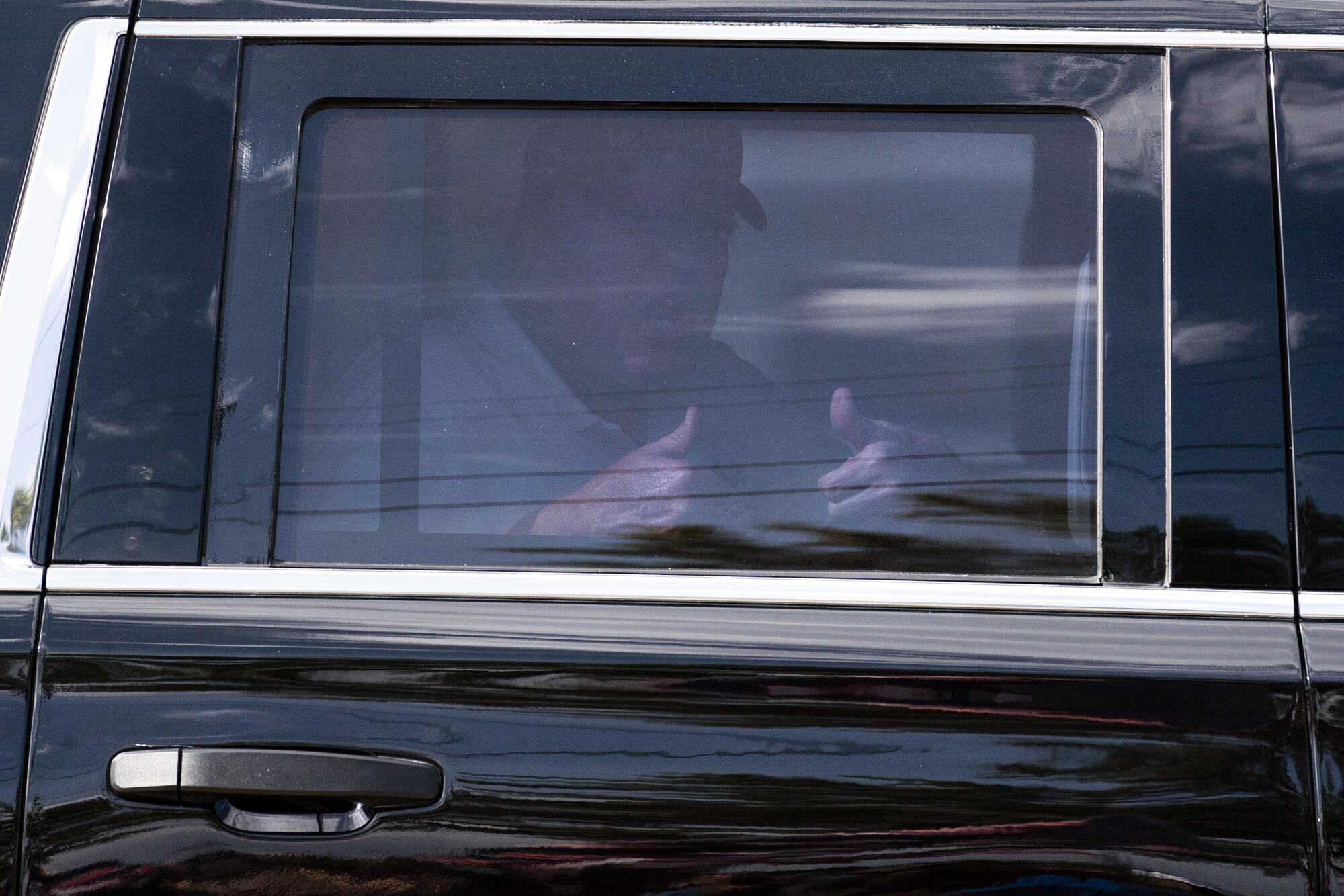 Donald Trump, wearing a red cap, gives two thumbs-up as he rides in the back of a car.