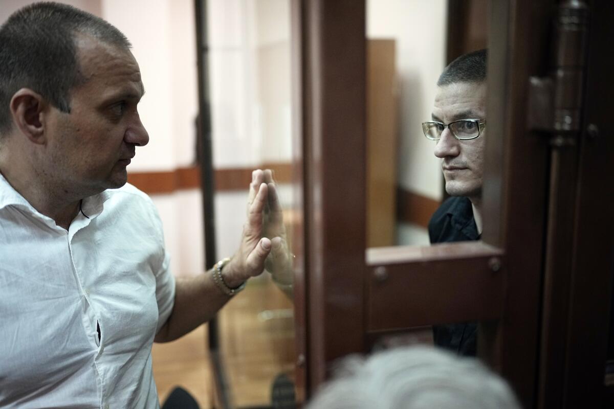 A man in a glass cage in court talks to another man on the other side of the glass. 