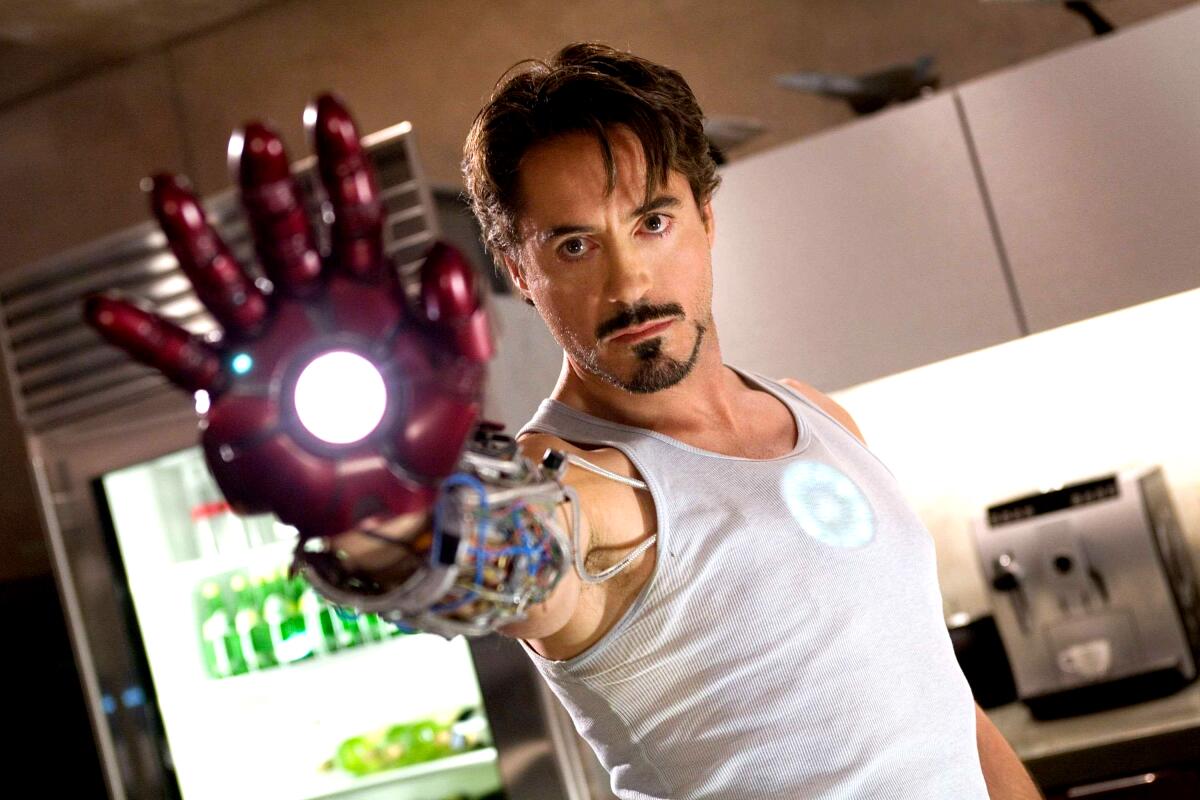 Robert Downey Jr., in a white tank top, reaches out his arm with his hand in a red Iron Man glove.