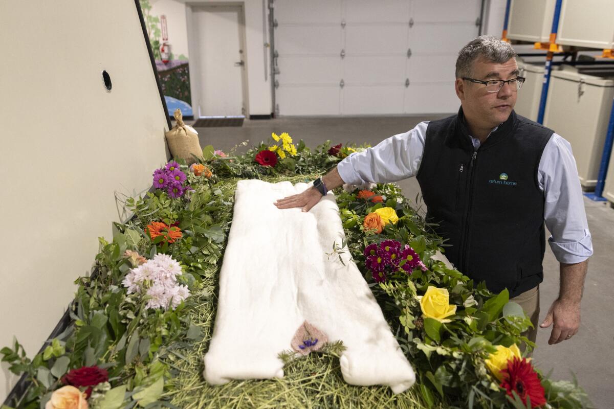 A man stands next to a human composting display that includes a shroud and flowers.