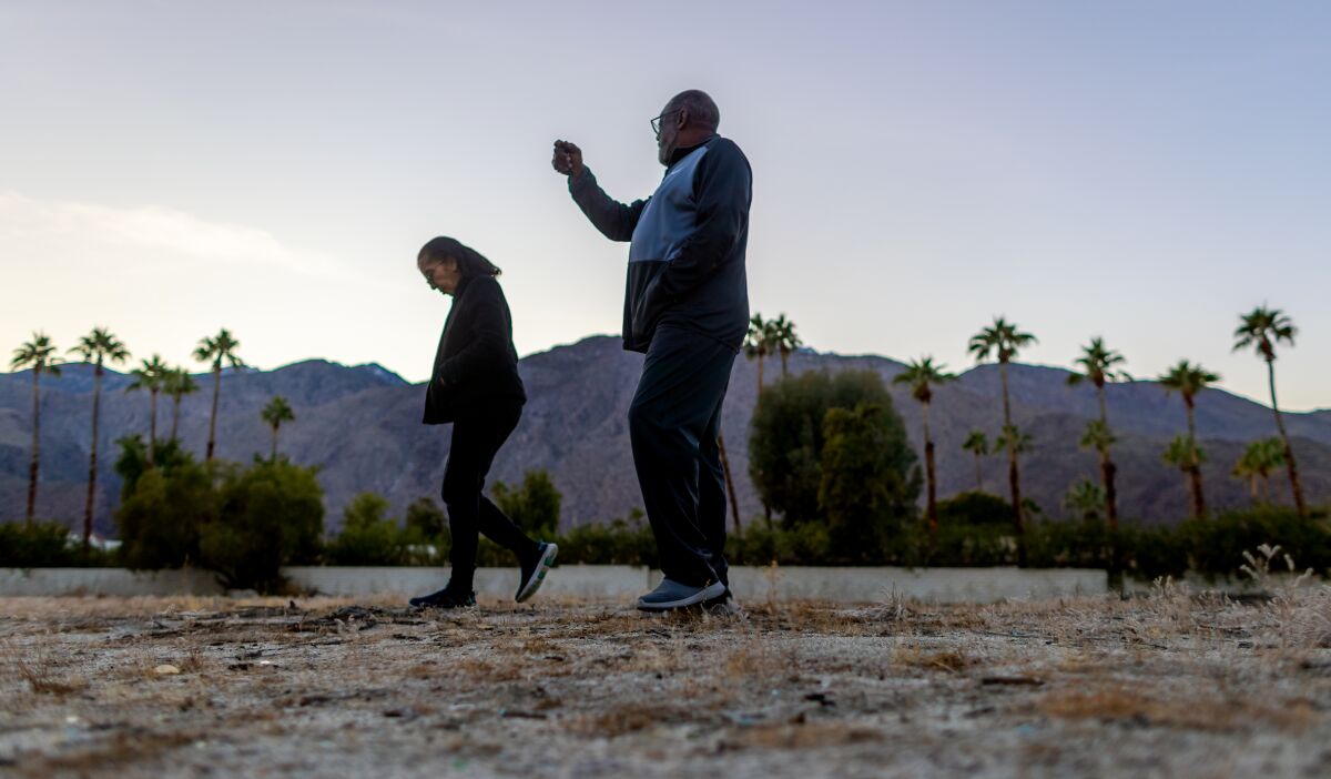 Two people walking through a vacant lot, with a row of palm trees and mountains in the background.