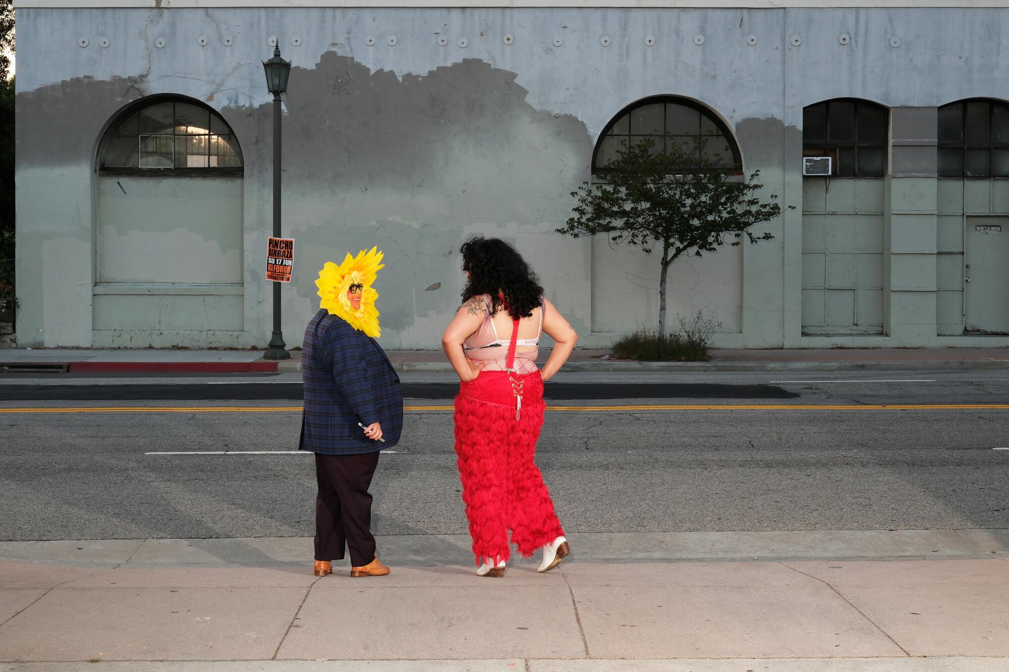 Seen from behind at a distance, a person in black suit and flower mask talks to a person in red on a sidewalk at dusk.