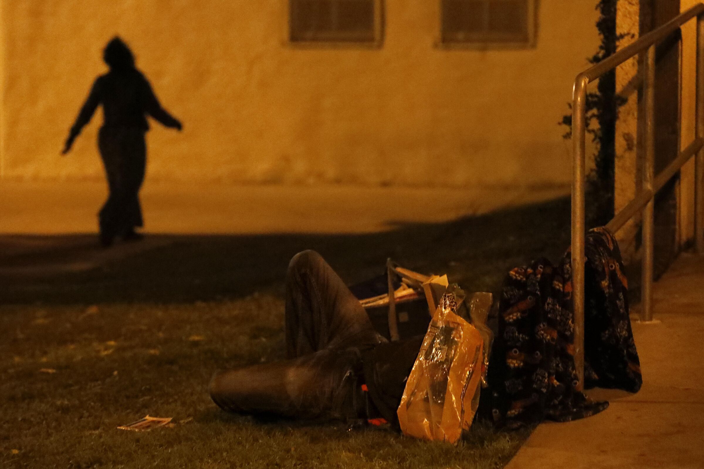 The shadow of a person appears near a person sleeping on the ground, obscured by personal belongings