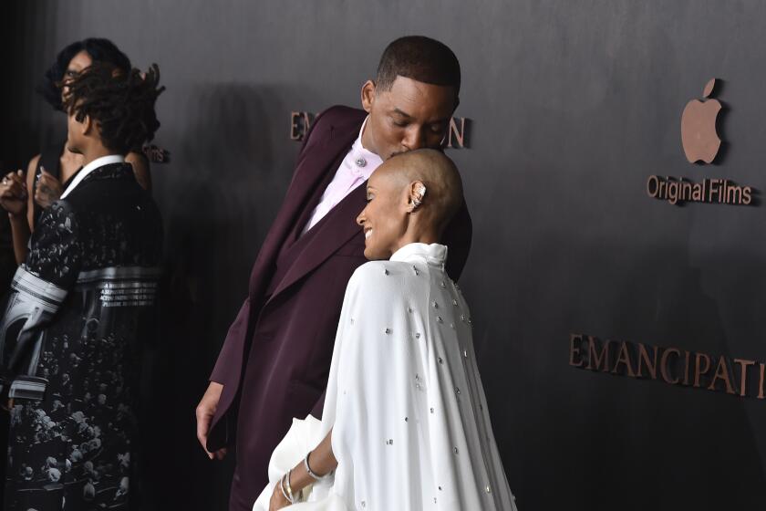 Will Smith in a burgundy suit leaning over to kiss Jada Pinkett Smith's shaved head. She's in a white gown and a cape