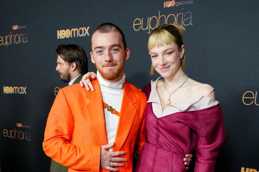 Angus Cloud is wearing a orange suit is posing next to Hunter Schafer who is wearing a maroon dress with white trim.