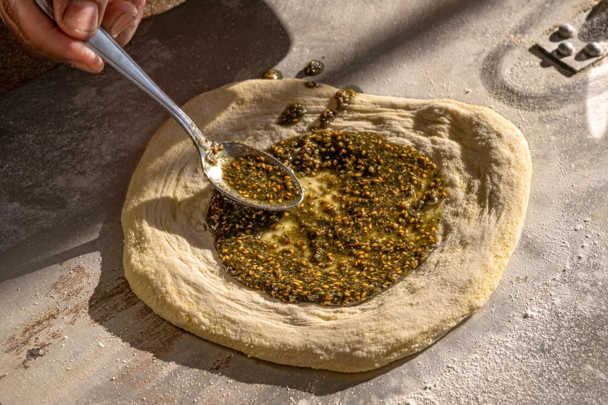 A hand holding a spoon, distributing a green sauce over a piece of round, flat dough. 