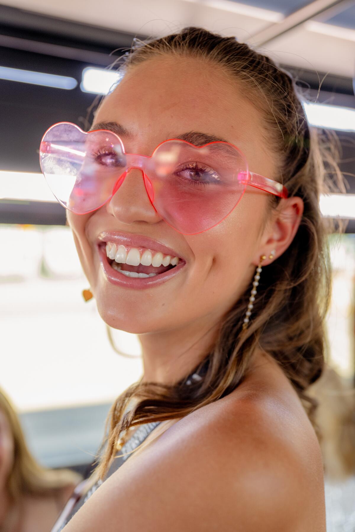 A young woman smiles, wearing pink hear-shaped sunglasses.