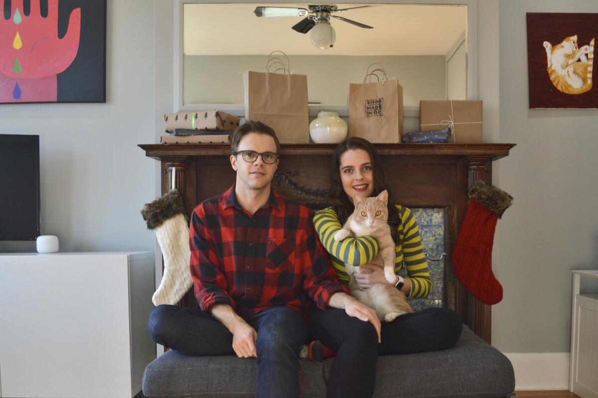 A man sits beside a woman, holding a cat, in front of a fireplace with Christmas stockings.