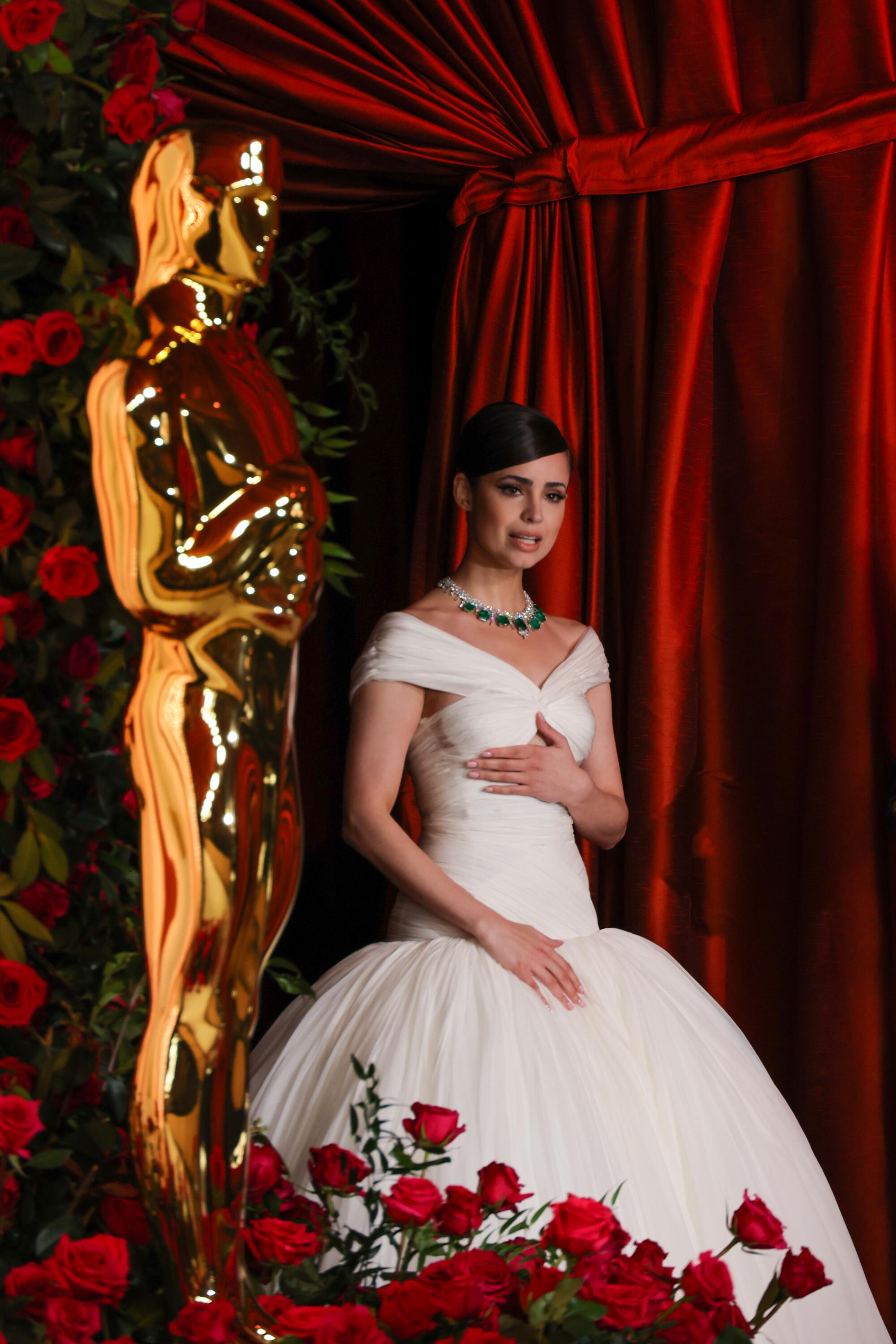 Sofia Carson in a white gown, standing next to a life-size Oscar statue.