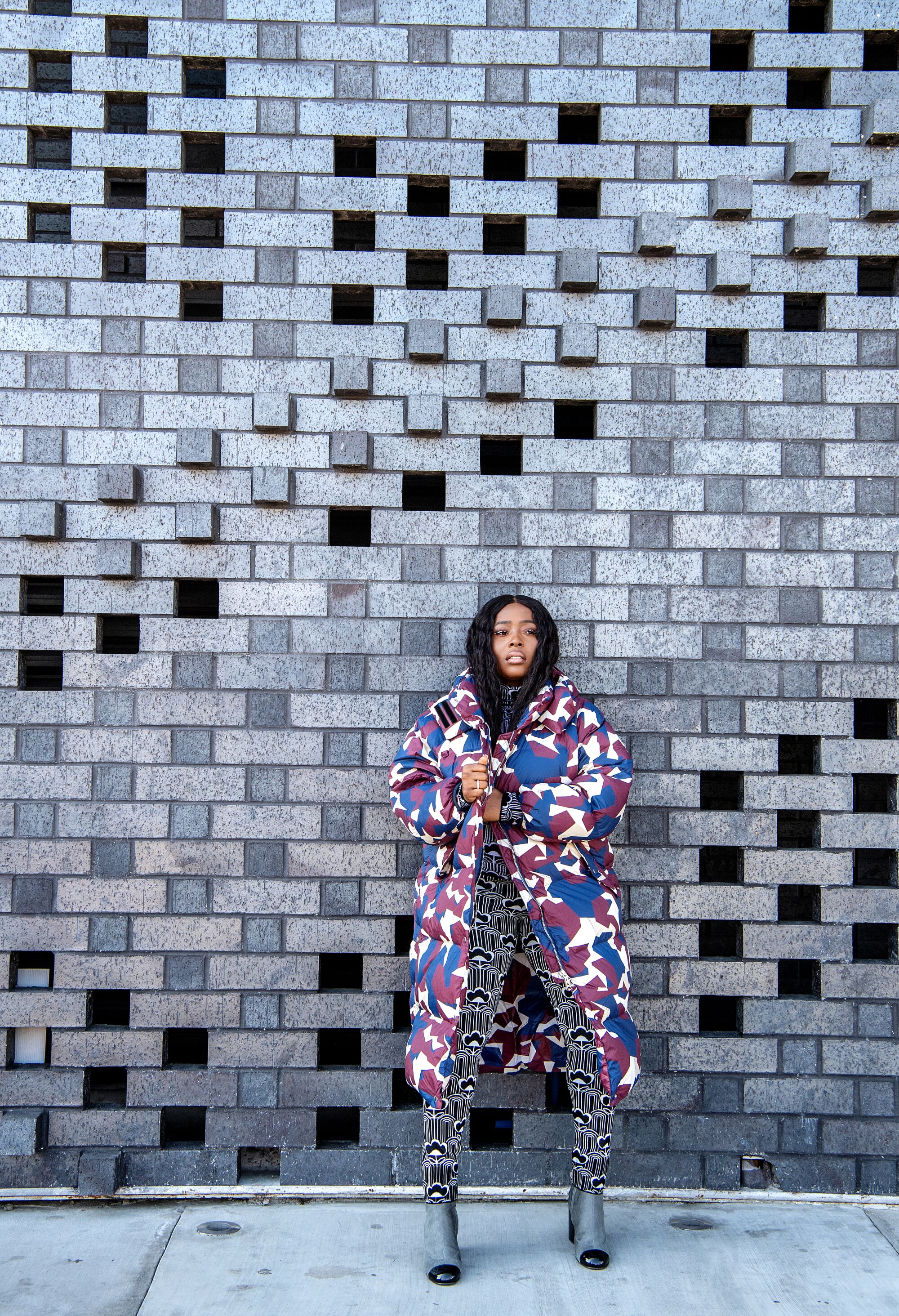 A woman in a colorful coat stands in front of a patterned wall.