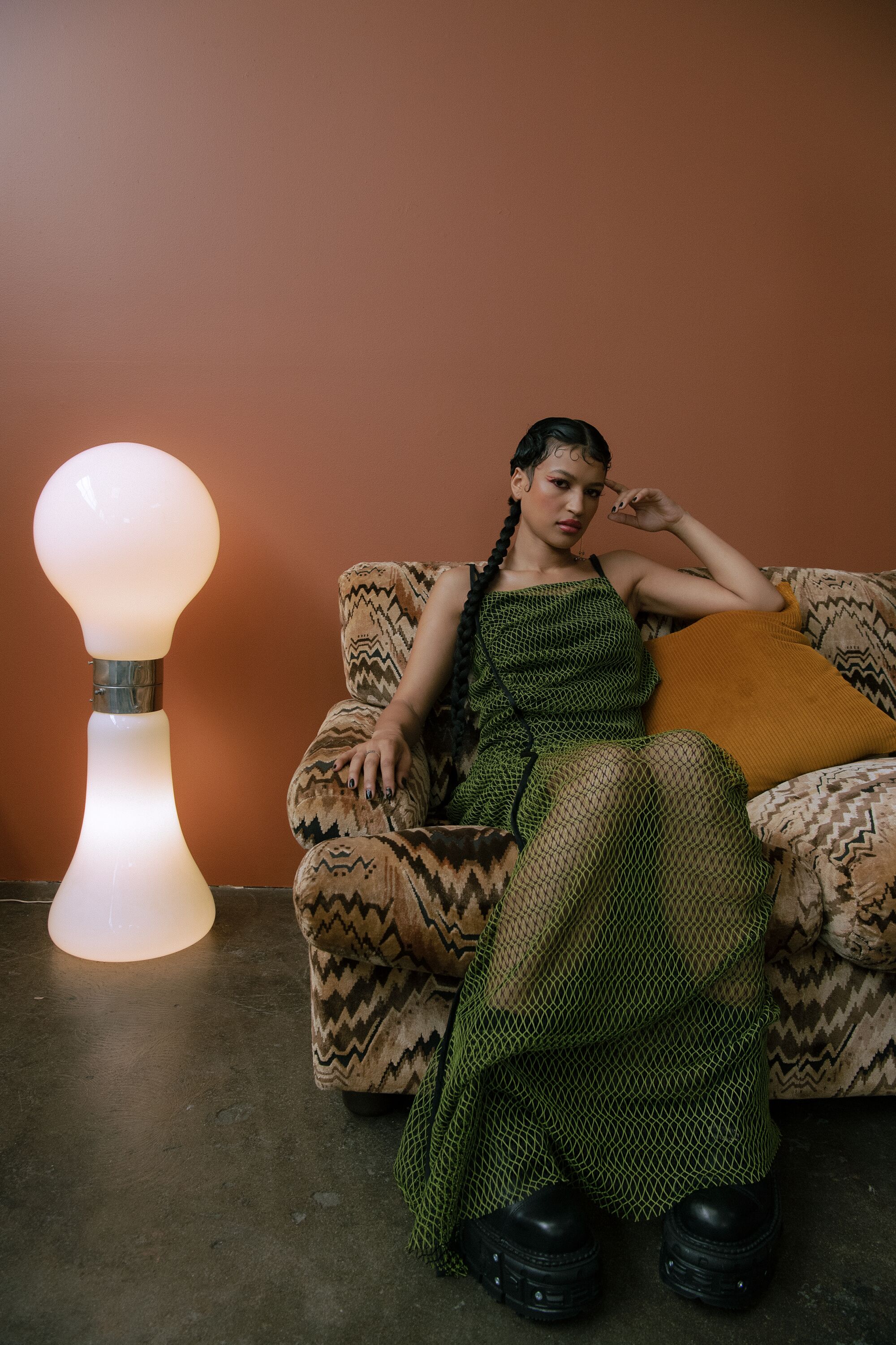 A model in a green dress lounges on a couch next to a glowing floor lamp.