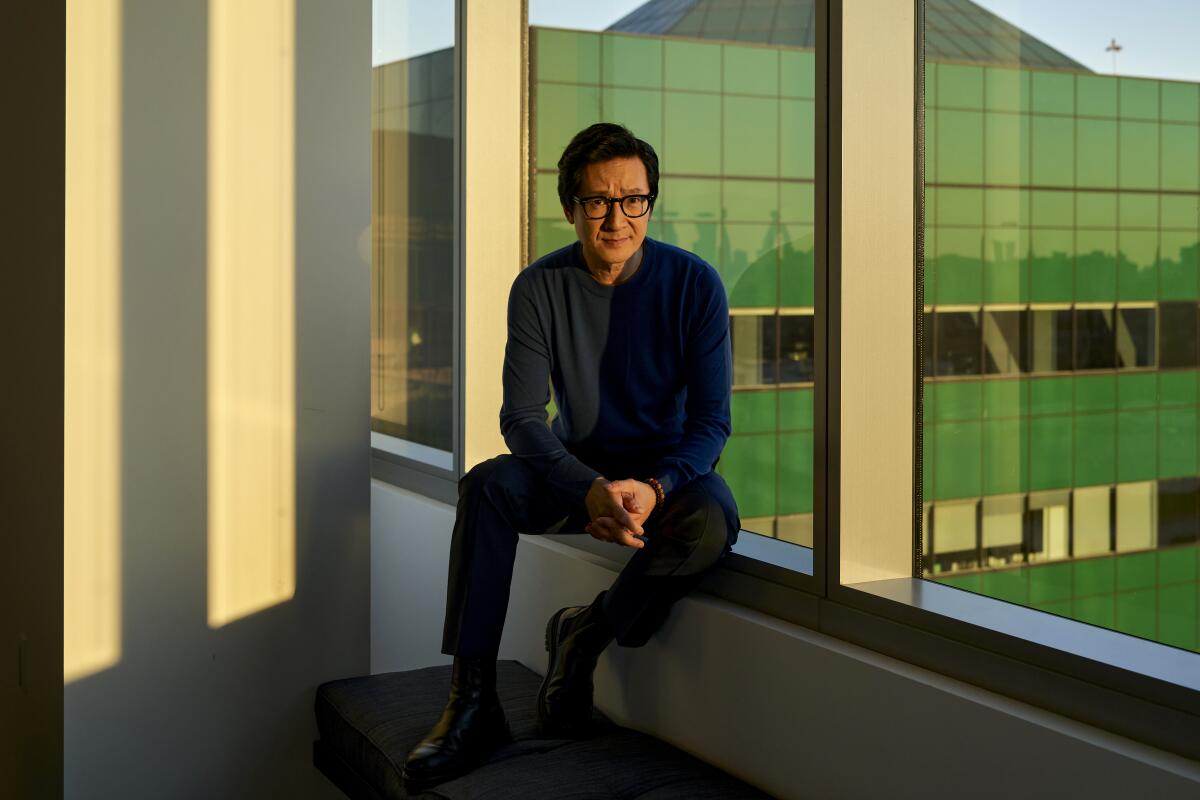 Ke Huy Quan sits on the inner ledge of a window for a portrait with a glass building in the backdrop.