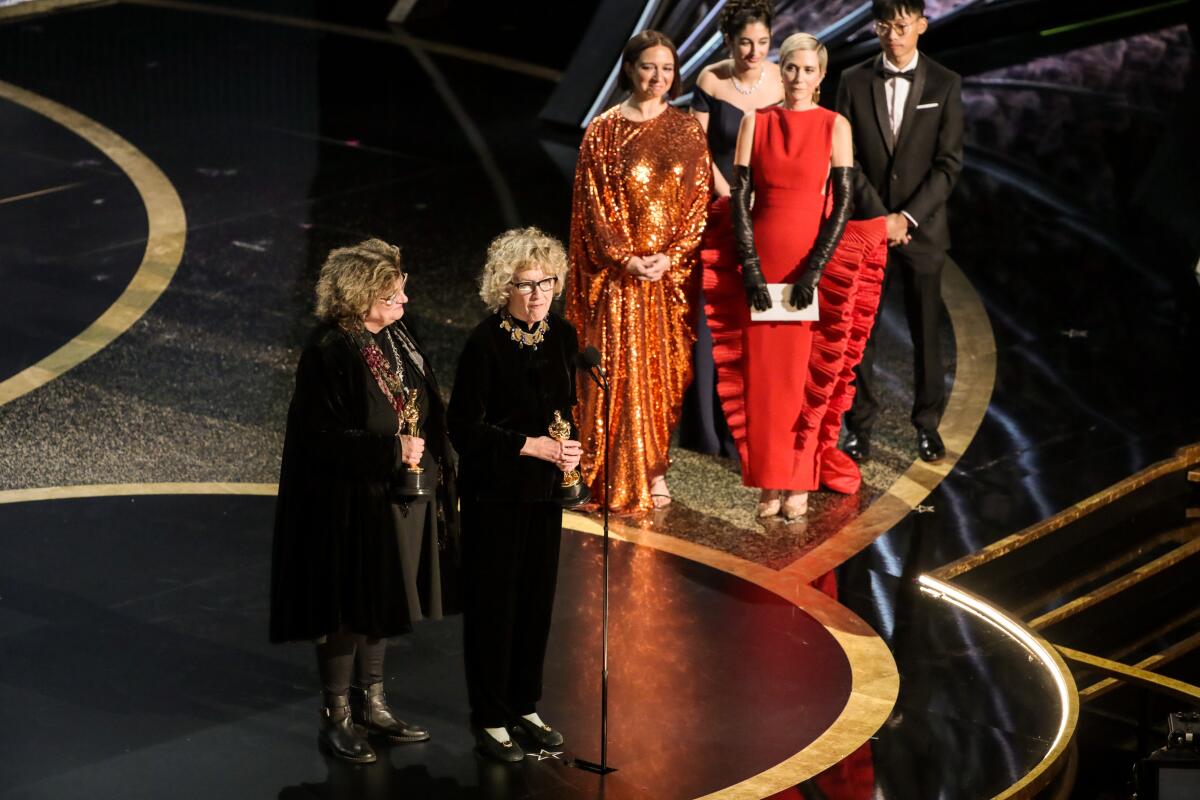 Two women in black dresses stand onstage at a microphone as other people stand nearby.