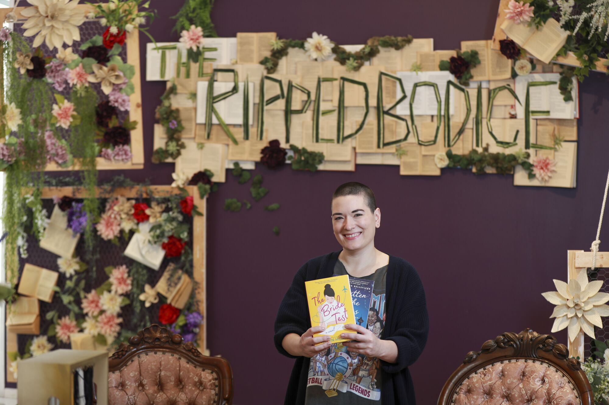 A woman with short hair holding a yellow book, surrounded by flowers and books on the wall.