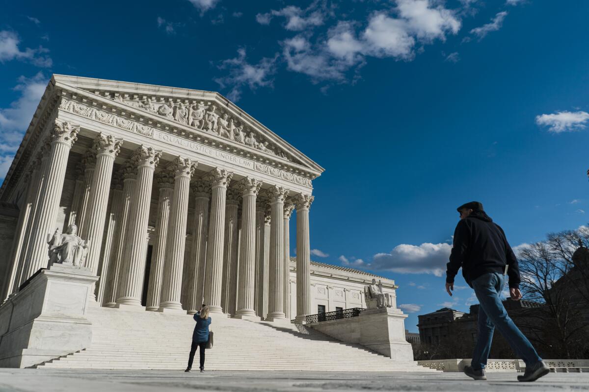 The Supreme Court building against a blue sky, as one person stands at the bottom of its stairway and another approaches.