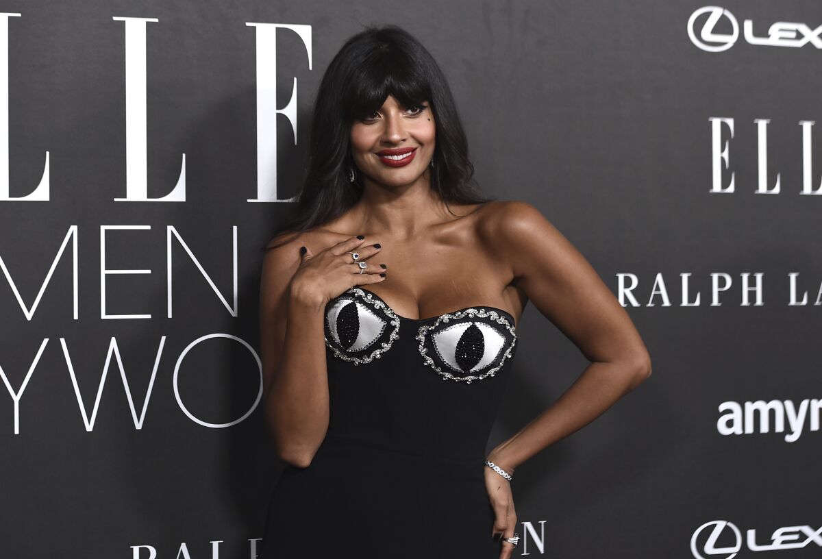 Jameela Jamil poses in a black dress with one hand on her hip and another at her collarbone