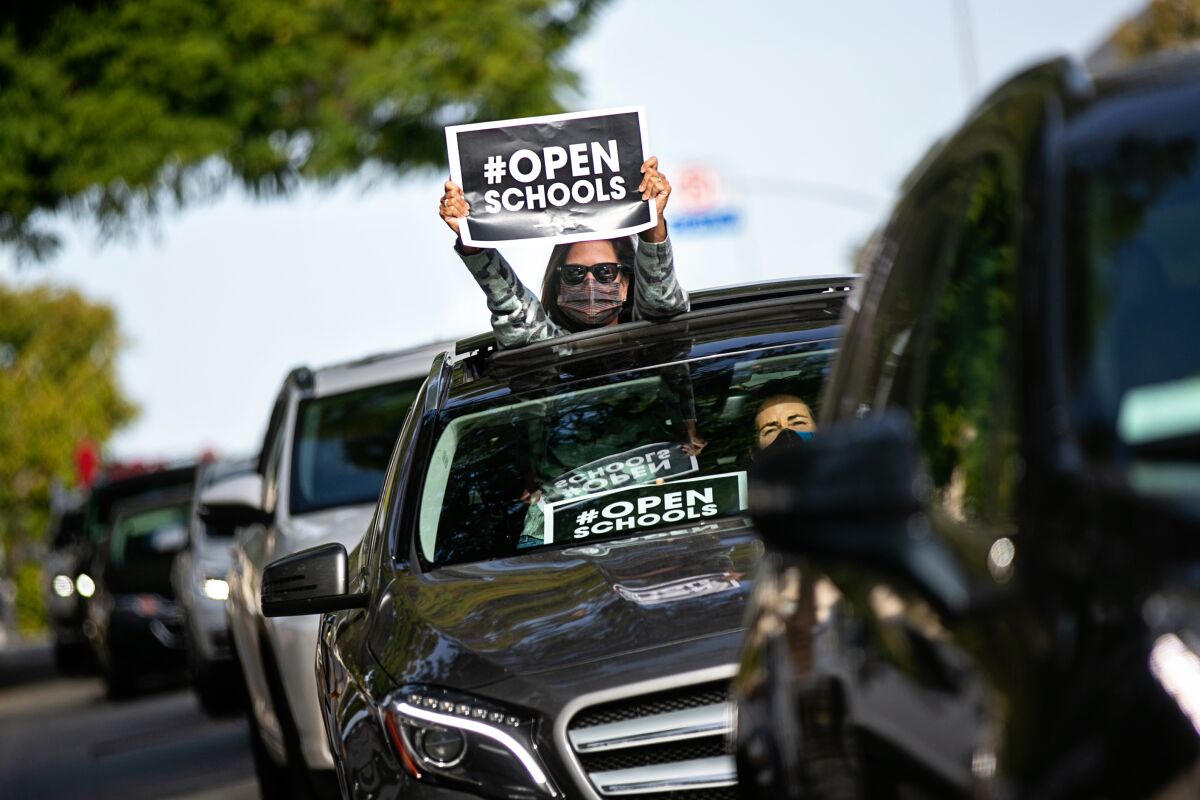 A woman stands out of a car's sunroof holding a sign that says Open Schools